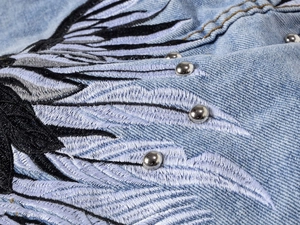 Detail of wing embroidery and silver metal studs on light blue denim vest for men