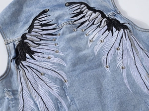 Detail of light blue denim vest with embroidered wing pattern and silver metal rivets