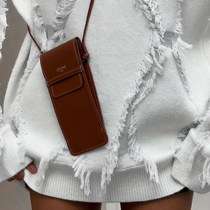 Close-up of a brown leather crossbody phone bag with brand logo over a white knit sweater