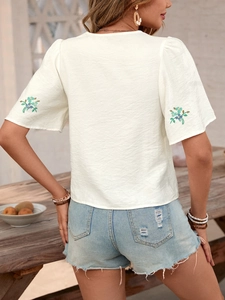 Detail shot of white linen shirt featuring teal floral embroidery on sleeves and denim shorts