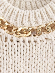 Detailed close-up of cream knit fabric featuring a chunky gold metal chain on the neckline