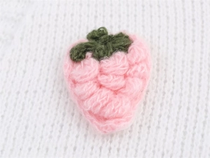 Nahaufnahme eines handgefertigten 3D-Häkel-Erdbeeranhängers auf rosa Strickjacke
