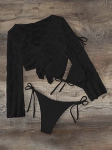 Flat lay of black three-piece bikini set with mesh long sleeve cover-up on wood floor