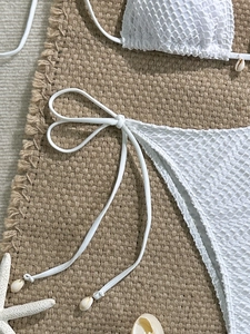 Detail of white mesh bikini with shell charms laid on a textured woven mat