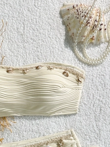 Dettaglio del top bikini bianco crema con texture a onde e ciondoli a forma di conchiglia