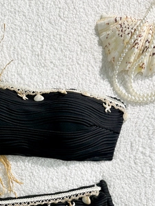 Bikini nero a coste con pizzo bianco e ciondoli a conchiglia su sfondo di morbido peluche
