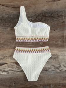 Flat lay of white one-shoulder bikini with colorful zigzag embroidery on wood background