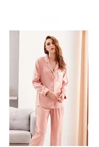 Model wearing pink satin silk pajama set with white piping and long sleeves in a home setting