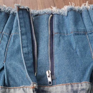 Detail view of denim waist with raw fringe edges, metal zipper, and orange contrast stitching