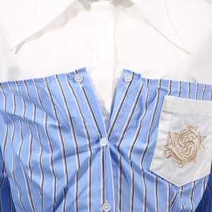 Dettaglio di camicia a righe blu con tasca bianca e ricamo dorato di lettere NSK sul petto