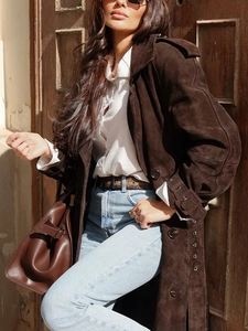 Elegant long dark brown suede trench coat with buttons worn by model against wood door