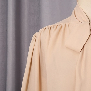 Detail of elegant beige chiffon blouse featuring a chic tie-bow neckline and pleated shoulders