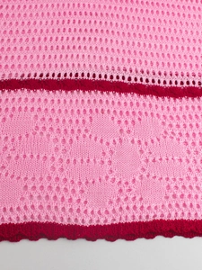 Detail of pink knitted fabric featuring burgundy heart crochet patterns and lace hem