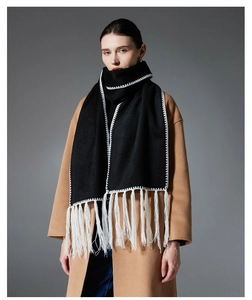 Dettaglio di una sciarpa nera con bordi a punto sopraggitto bianchi e lunghe frange su un cappotto cammello