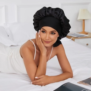 Silky black satin sleep cap with wide elastic headband worn by a model in a cozy bedroom