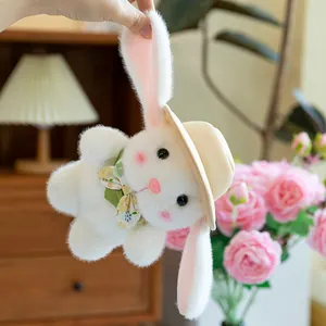 Cute white plush bunny with straw hat and floral bowtie held by ears in a cozy setting