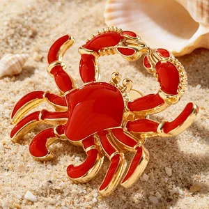Detailaufnahme einer roten Emaille-Krabbenbrosche auf goldenem Sand am Strand.