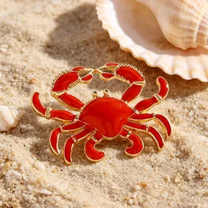 Detaillierte Makroansicht einer roten Emaille-Krabbenbrosche auf Strandsand mit Muscheln