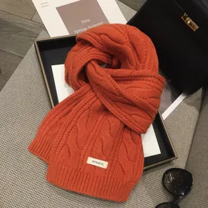 Detail showing orange wool scarf with chunky cable knit texture and 100% wool label