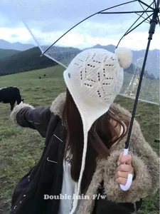 Outdoor shot of a white wool knit trapper hat featuring hollow patterns and pompom