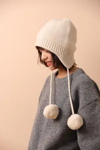 Detalle del gorro de lana crema con orejeras, pompones suaves y textura de punto acanalado
