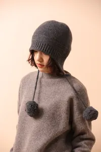 Primer plano de gorro de lana gris oscuro con pompones decorativos y correas laterales