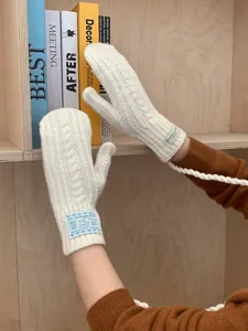 Detail of chunky cable knit cream mittens featuring decorative labels and neck lanyard