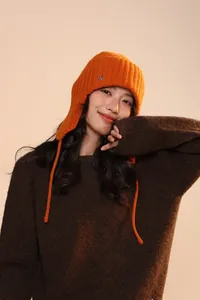 Dettaglio del cappello in maglia arancione con paraorecchie, logo in metallo e morbida trama