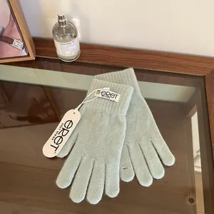 Detail view of sage green knit gloves with m·ERET label on a glass tabletop