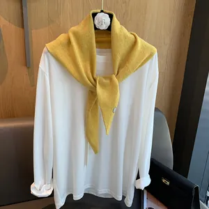 Detail eines gelben Wollstrickschals mit Metallemblem auf weißem Langarmshirt