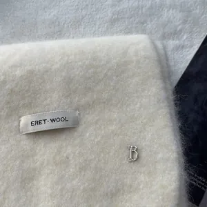 Macro detail of soft white wool knit scarf featuring ERET WOOL label and silver metal B charm