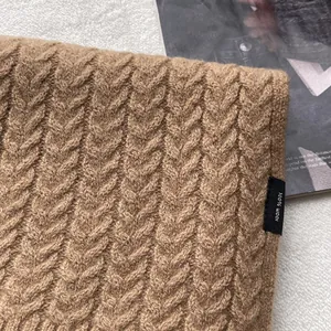 Dettaglio di una sciarpa di lana a trecce color cammello con etichetta 100% lana