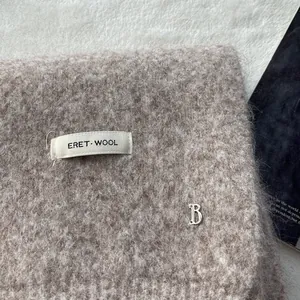 Close-up of soft oatmeal wool knit scarf with premium metallic B pendant and brand label