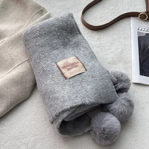 Thick grey knit scarf with decorative pom-poms and brand tag on soft texture background
