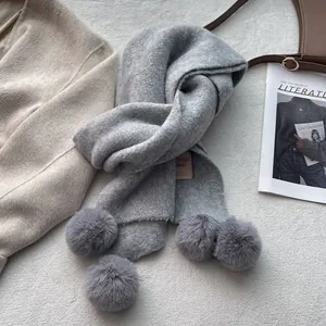 Detailed close-up of a grey knit scarf featuring large decorative fur pom-poms and fabric label
