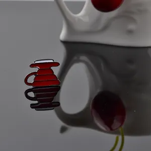 Red enamel drip coffee pot lapel pin on black reflective surface with cherry decor