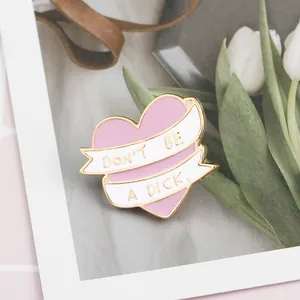 Close-up of heart enamel pin with "DON'T BE A DICK" slogan on pink checkered background