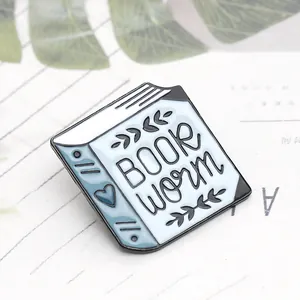 Dettaglio della spilla smaltata Book Worm con cuore blu su uno sfondo di foglie verdi
