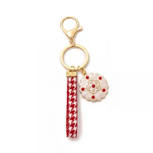 Detail of a golden keychain with a white enamel flower and red houndstooth ribbon strap