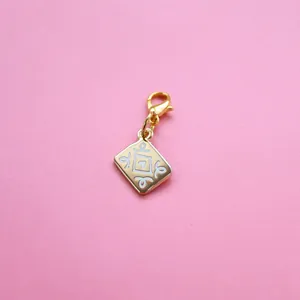 Close-up of golden metal charm with white enamel pattern and lobster clasp on pink backdrop