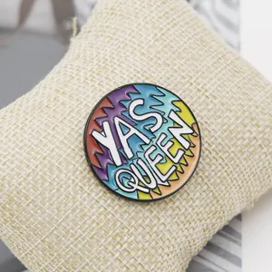 Close-up of a colorful enamel brooch with Yas Queen text on a beige fabric cushion