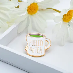 Close-up of 'FUELED by TEA' white enamel mug brooch with floral background