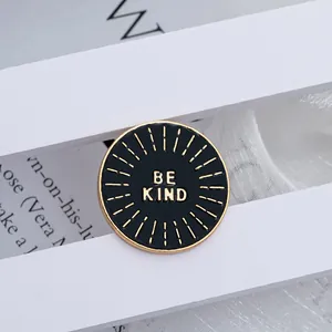 Detail shot of a round black enamel BE KIND brooch with gold sunburst on white fabric