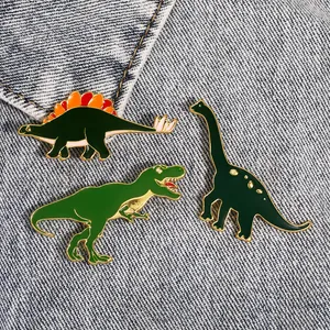 Detailansicht von drei süßen Dinosaurier-Emaille-Pins auf blauem Denim-Hintergrund