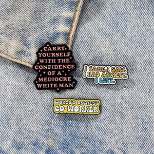 Close-up of funny quote enamel pins in black, white, and yellow on blue denim fabric