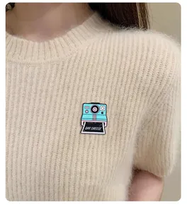 Dettaglio della spilla smaltata a fotocamera vintage con scritta Say Cheese su maglione