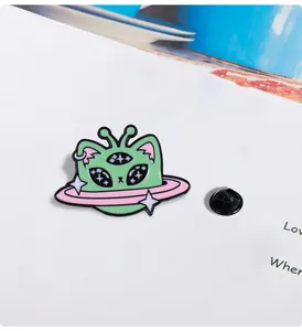 Close-up of a green alien cat enamel pin with three eyes and pink Saturn ring