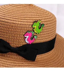 Close-up of green and pink dragon enamel pins on a straw hat with a black bow ribbon