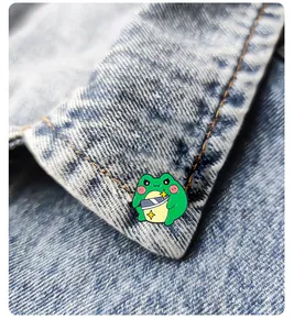 Detail of a funny cartoon green frog enamel pin holding a knife on denim fabric