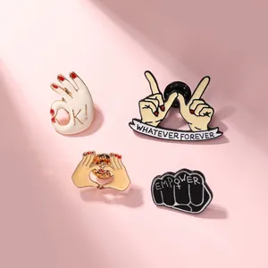 Flat lay of enamel hand gesture pins including OK and Empower on a pink surface